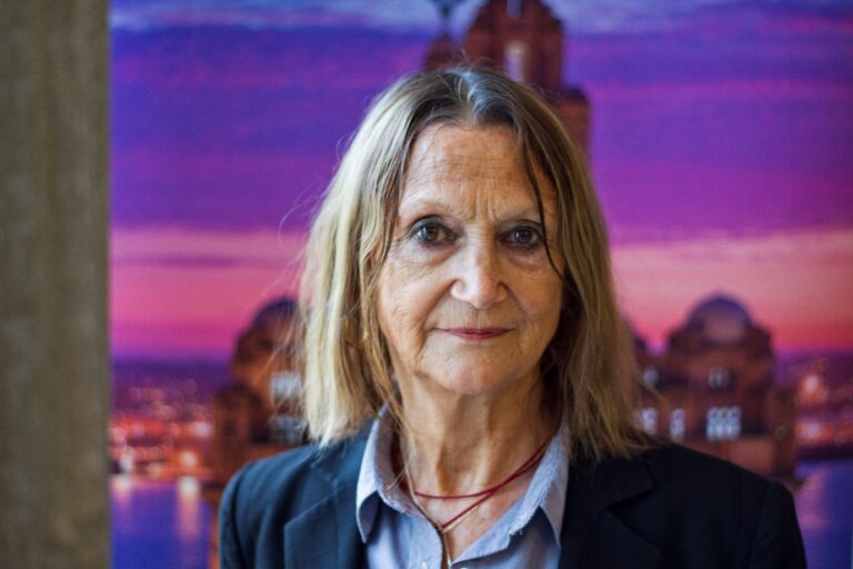 a photo of julia baird in present day behind a pink and purple sunset liverpool city backdrop